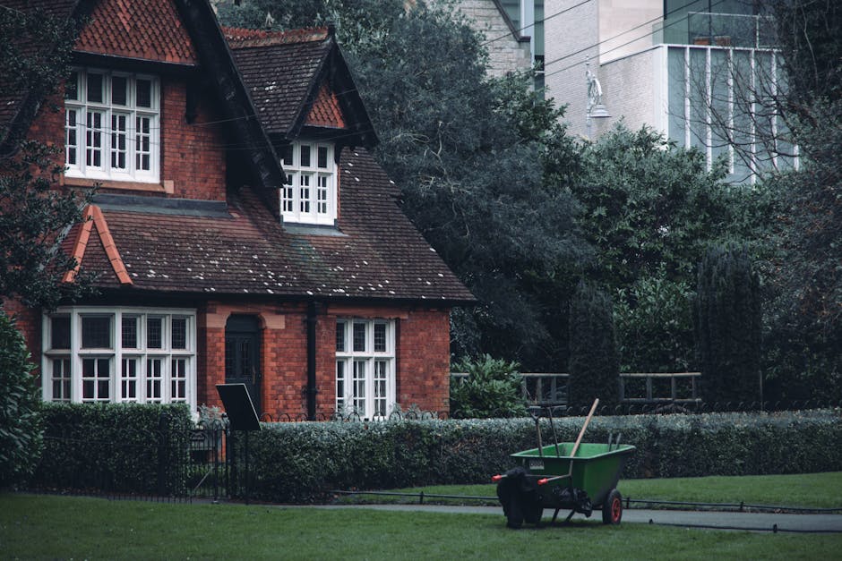 A red-brick cottage with white-framed bay windows and a steeply pitched roof covered in dark tiles, situated within a well-maintained garden. The garden features a neatly trimmed hedge border, lush gr